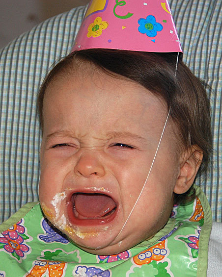 baby with food on face, in party hat, crying with wide open mouth