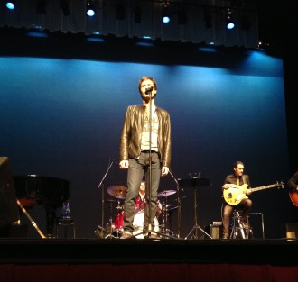 Eric McCormack onstage in a Stand Up 2 Cancer shirt, singing a song at his concert