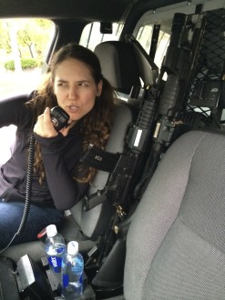 Aurora De Lucia giving a semi-tough face into a walkie-talkie in the front of a police cruiser