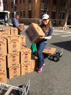 Aurora De Lucia smiling and holding a box of food while working with Food Forward at a Farmers' Market